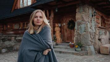 Blonde woman wrapped in gray blanket enjoying alpine view near cozy wooden house, fresh air and mountain stillness filling calm atmosphere. Stillness flows in scenery, stillness brings mindful pause video
