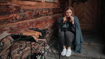 Cozy caucasian woman drinking hot beverage, sitting wrapped in blanket on rustic cabin porch, enjoying tranquil scenery with serene relaxation. Woman taking sip of hot drink, looking away and enjoying video