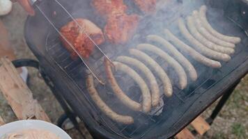 Close up sizzling smoke rising from juicy sausages and meat grilling on barbecue, professional chef using tongs for even roasting and perfect caramelization. Chef turns over grilling meat with tongs video
