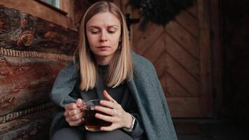 Woman with distant gaze holds warm mug near face, lost in thoughts on cabin porch, woman absorbs quiet morning stillness, woman soft expression conveys peaceful reflection. Slow Living Concept video