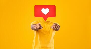 A person wearing a yellow shirt points toward the viewer in a vibrant setting filled with a bright yellow background. Instead their head is heart icon symbolizing likes and affection in social media. photo