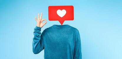 A person dressed in a blue sweater makes an OK hand gesture, with a heart icon superimposed above where their head would be. The simple blue backdrop highlights the playful social media theme. photo