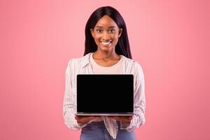 Smiling African American woman showing laptop with empty screen on pink studio background, mockup for website design. Lovely black lady with modern gadget, blank space. Online life concept photo