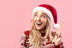 Excited millennial lady in Christmas outfit writing New Year resolutions in copybook on pink studio background, banner design with empty space. Cheerful young woman making Xmas wish list photo