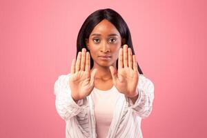 Millennial black woman gesturing STOP with both hands on pink studio background. Assertive African American lady saying NO, showing rejection gesture, expressing refusal or denial photo