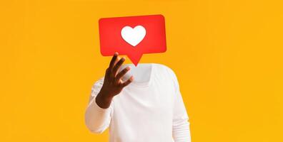 A person wearing a white long-sleeve shirt outstretching hand with large red heart icon instead of his head, bright yellow backdrop. This represents engagement on social media platforms. photo