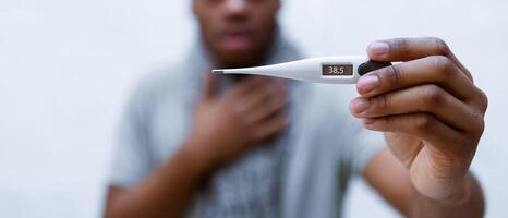 Fever. Shocked ill african american man showing thermometer with high temperature result to camera standing over gray background. Coronavirus, influenza symptom concept. Shallow depth, selective focus photo