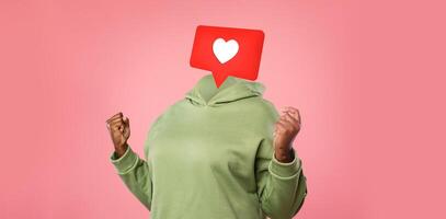 A person in a green hoodie expresses excitement and joy as a heart icon hovers instead of their head. Background is a soft pink, emphasizing the emotion linked to social media interactions and likes. photo