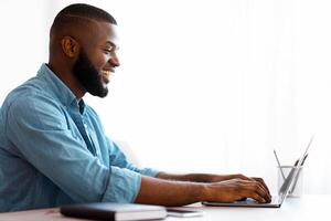 Black Young Man Using Laptop At Home, Enjoying Remote Work. Smiling African American Freelancer Guy Using Computer For Online Job, Typing On Keyboard And Looking At Screen, Side View With Free Space photo