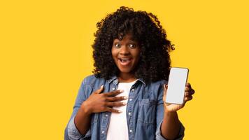 Amazed young black lady showing modern smartphone with empty screen, sharing her positive emotions after using nice mobile application, yellow studio background, copy space photo