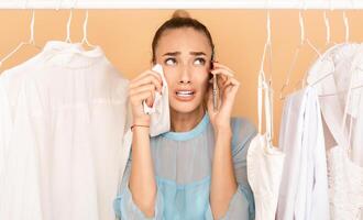 I Have Nothing To Wear. Portrait of upset stressed woman talking on mobile phone and crying, wiping tears with shirt, having panic, standing in rack of clothes between hangers photo