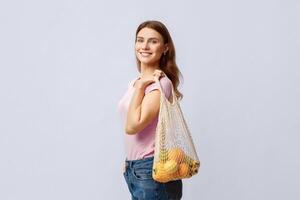 Zero Waste Concept. Young Woman Holding Mesh Eco Bag With Organic Fruits And Vegetables, Using Reusable Crochet Net Bag For Grecery Shopping, Standing Over Light Gray Studio Background, Copy Space photo