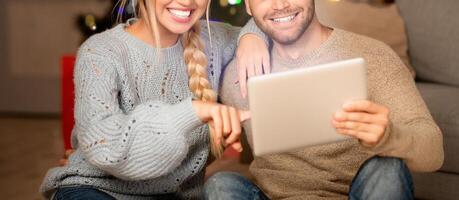 Loving couple having fun together at home on xmas, sitting on floor by couch and using digital tablet, copy space. Emotional young man and woman playing game online on pad together photo