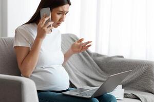 Stressed Pregnant Woman Working Remotely With Laptop Computer And Talking On Cellphone At Home, Concerned Expectant Lady Suffering Business Problems, Sitting On Couch In Living Room, Copy Space photo