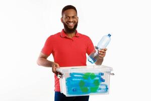 African Man Putting Plastic Bottle To Recycle Box Posing Smiling To Camera On White Studio Background. Used Plastic Disposal And Waste Sorting, Junk Recycling Concept photo