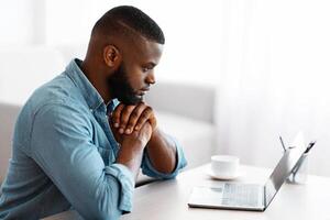 Pensive African American Man Working With Laptop At Home Office. Black Freelancer Guy Sitting At Desk And Looking At Computer Screen With Thoughtful Face Expression, Side View With Copy Space photo