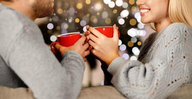 Closeup of smiling woman and unrecognizable man sitting on couch, drinking cocoa, copy space. Happy man and woman enjoying Christmas together at home, sitting on sofa over shining xmas tree, side view photo