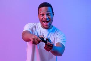 Overjoyed Black Gamer Man With Joystick Playing Games In Neon Lighting, Emotional Excited Guy Having Fun While Standing In Neon Lighting Over Purple Studio Background, Looking At Camera photo
