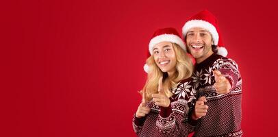 Portrait of cheerful young couple in Christmas sweaters pointing at camera over red background photo