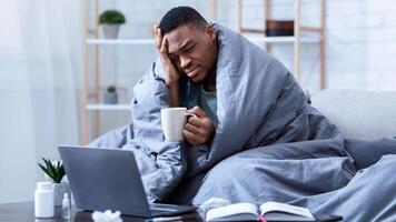 Sick African American Man Using Laptop Working While Being Ill, Having Fever And Headache Sitting On Sofa Wrapped In Blanket At Home. Guy Browsing Internet For Influenza, Cold, And Covid-19 Symptoms photo