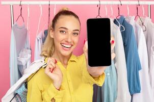 Recommending Mobile App. Portrait Of Cheerful Stylish Lady Showing To Camera Mobile Phone With Black Empty Screen For Mockup, Holding Hanger With Clothes, Posing With Rack In The Pink Background photo