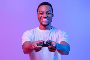 Modern Entertainment. Portraif Of Handsome Smiling Black Guy In Neon Light With Joystick In Hands Looking At Camera, African Man Playing Games, Standing Over Purple Background, Copy Space photo