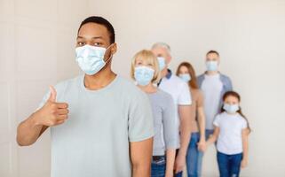 Black guy in face mask waiting in queue for coronavirus vaccination at clinic, showing thumb up gesture, copy space. Panorama. Covid-19 immunization, protection against global virus concept photo