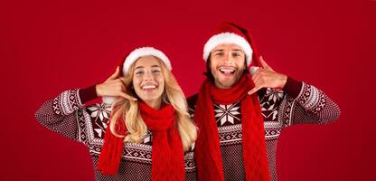 Joyful young couple in Christmas sweaters and Santa hats showing CALL ME gesture on red background photo