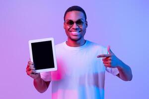 Cheerful Black Man Pointing At Blank Digital Tablet Screen, Demonstrating Copy Space For Your App Or Website Design, Standing Under Vivid Neon Light Over Purple Studio Background, Mockup Image photo