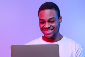 Handsome Smiling Black Programmer Guy Working Online On Laptop In Neon Light, Cheerful African American Freelancer Man Using Computer In Neon Light Over Purple Studio Background, Closeup Portrait photo