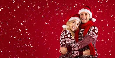 It's Christmas. Beautiful romantic couple in Santa hats and warm sweaters hugging on red background photo