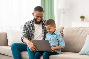 African American Young Father And Son Using Laptop Together, Dad Teaching His Kid To Use Computer And Browsing Internet Sitting On Sofa At Home. Family Lifestyle, Technology Concept photo
