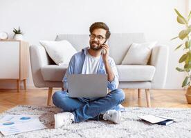 Arab Freelancer Guy Using Laptop And Talking On Cellphone At Home, Smiling Eastern Man In Eyeglasses Working Remotely With Computer And Documents While Sitting On Floor In Living Room, Copy Space photo