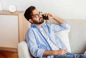 Handsome Eastern Man Talking On Cellphone While Relaxing On Sofa At Home, Millennial Arab Guy Speaking On Phone With Friends, Sitting On Couch In Living Room, Enjoying Pleasant Conversation, Side View photo