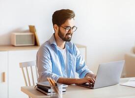 Millennial arab freelancer man working online on laptop at home office, eastern guy sitting at desk looking at computer screen and typing on keyboard, enjoying distant job opportunities, copy space photo