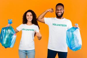 Volunteers Team. Multiracial Man And Woman Holding Blue Garbage Bags With Wasted Plastic Posing In Studio Over Yellow Background. Keep Environment Clean, Ecology And Volunteering Job photo