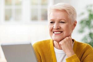 Closeup of happy old grey-haired woman using digital tablet in kitchen, empty space. Happy modern grandmother reading culinary blog or book online, surfing on Internet on newest pad at home photo
