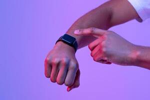Smart devices. Unrecognizable african man pressing button on smart watch with black screen on his hand, standing in neon lighting over purple studio background, cropped image with free space, mockup photo