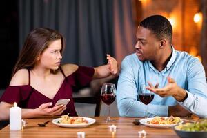 Annoyed Black Guy Blaming His Girlfriend For Using Smartphone During Romantic Dinner In Restaurant, Young Woman Showing Stop Gesture With Hand, Multicultural Couple Having Relationship Problems photo