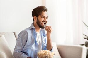 Cheerful eastern man spending free time at home, watching TV and eating popcorn. Happy arab guy enjoying comedy movie while relaxing on couch in living room, side view with free space photo