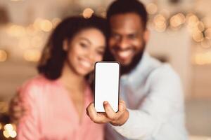 People And Technology Concept. Smiling African American Man Holding And Showing Smartphone With White Blank Empty Screen For Mockup, Hugging Woman In Blurred Background, Selective Focus photo