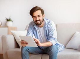 Handsome Young Arab Man Using Digital Tablet Computer At Home, Millennial Eastern Guy Browsing Internet Or Checking Social Networks While Relaxing On Couch And Smiling At Camera, Free Space photo