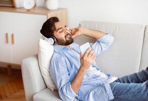 Eastern man listening music on smartphone online with wireless headphones, relaxing on couch at home, millennial arab guy enjoying his favorite playlist while lying on sofa with cellphone, side view photo