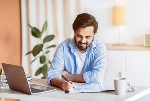 Smiling Arab Man Working With Documents And Laptop At Home Office, Handsome Eastern Male Entrepreneur Sitting At Desk With Computer, Checking Financial Reports And Taking Notes, Free Space photo