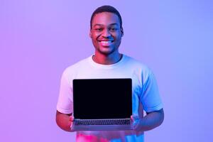 New Website. Happy African American Man Holding Laptop Computer With Black Blank Screen, Showing Free Copy Place For Your Mockup Design, Standing In Neon Lighting Over Purple Background photo