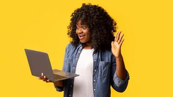 Cute young black lady waving and smiling at laptop screen, having call over yellow studio background, copy space. Happy african american woman having online party with friends photo
