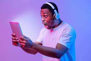 Portrait Of Amazed Black Guy In Wireless Headphones Looking At Glowing Screen On Digital Tablet With Excitement, Emotionally Reacting To New Game Or App, Standing In Neon Light Over Purple Background photo