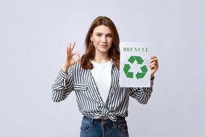 Smiling Young Lady Holding Motivational Placard With Recycle Sign And Showing Ok Gesture, Agitating To Save Ecology And Nature With Zero Waste Concept, Standing Over Grey Studio Background, Free Space photo