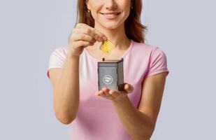 A millennial woman happily participates in recycling by placing a yellow item into a small recycling bin. She showcases her commitment to eco-friendly practices in a minimalistic background. photo