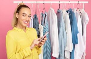 Fashion And Style. Portrait of smiling woman stylist holding and using smartphone, texting sms or making purchases in online store standing near rack with clothes over pastel pink studio background photo